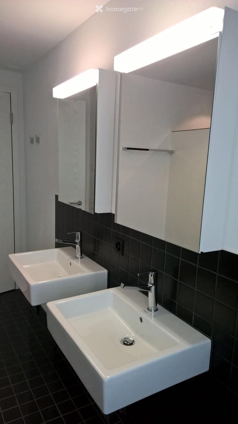 Modern bathroom with white vanity sinks, black tile walls, and recessed lighting above the mirrors