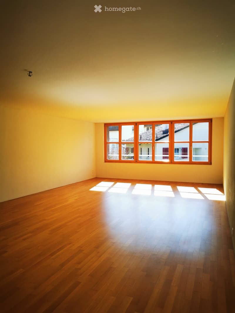 Spacious living room with large windows, hardwood floors, and warm yellow walls. The room is filled with natural light from the windows, creating a bright and airy atmosphere.