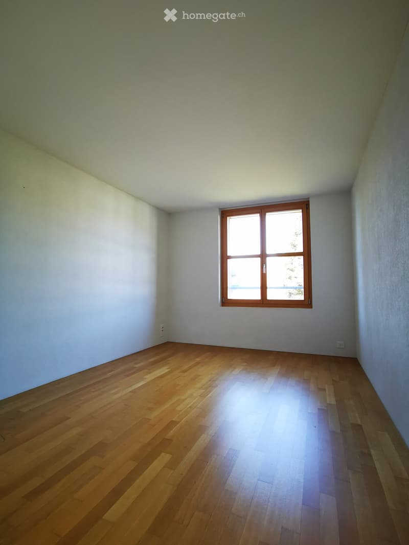 The image shows a spacious bedroom with white walls and a large window. The room has a hardwood floor and appears to be empty, ready for furnishing.