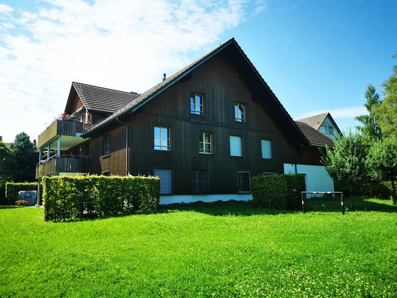 2-story wooden house with dark exterior, balconies, large windows, surrounded by a well-maintained lawn and garden