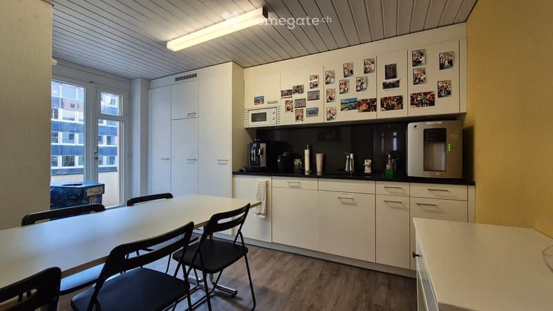 The image shows a modern kitchen with white cabinets, a black countertop, and various kitchen appliances such as a microwave and a dishwasher. The kitchen also has a dining table with black chairs. The walls are decorated with framed photos, and the ceili