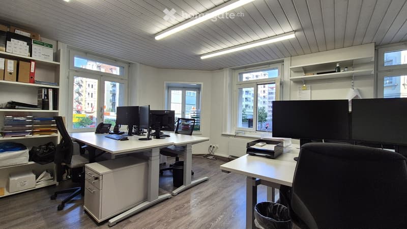 The image shows a modern office space with a large desk, multiple computer monitors, shelves, and storage units. The room has large windows providing natural light, and the ceiling is lined with fluorescent lighting fixtures. The floor appears to be a lam