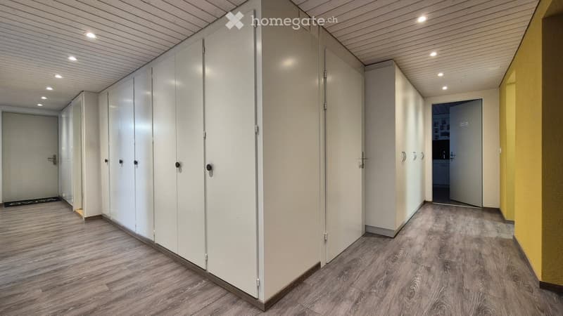 The image shows a locker room with multiple white lockers lining the walls. The floor is made of a gray wood-like material. The room is well-lit with recessed lighting in the ceiling.