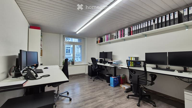 Spacious office with multiple workstations, computer monitors, and storage shelves. The room has a large window, hardwood flooring, and a modern, organized layout.