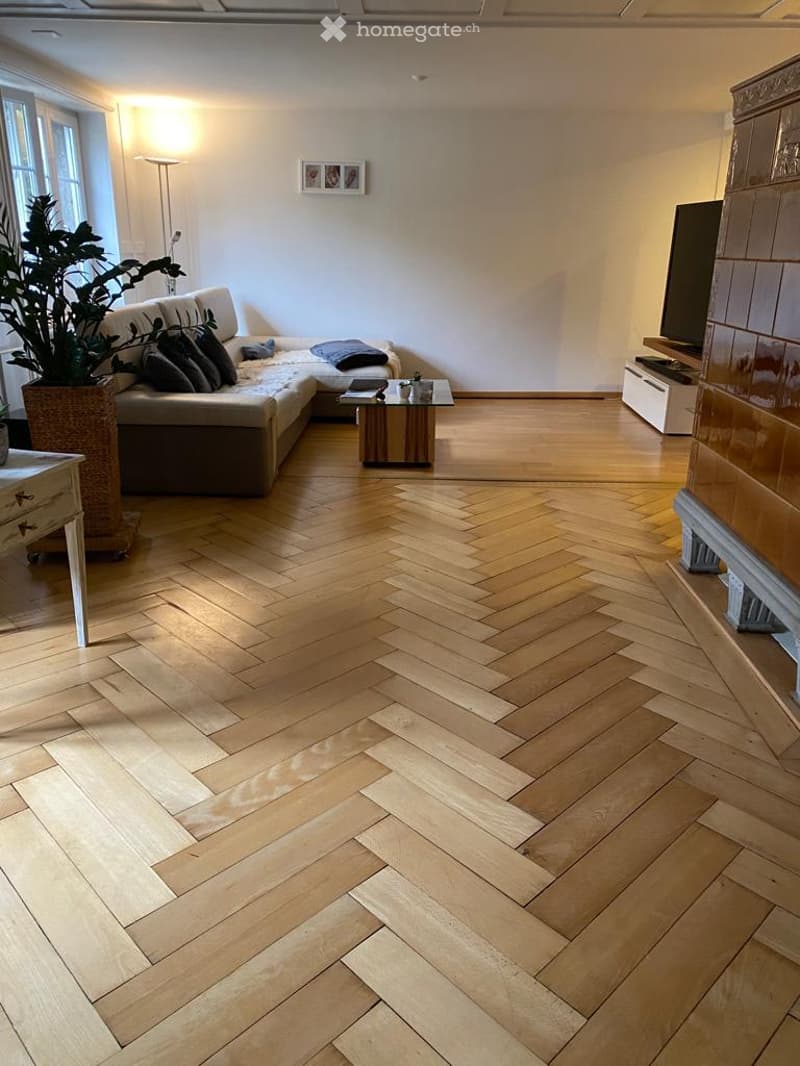 A room with wooden herringbone floor, white walls, a couch with pillows, a table, a lamp, a potted plant, a TV, and a picture frame on the wall.