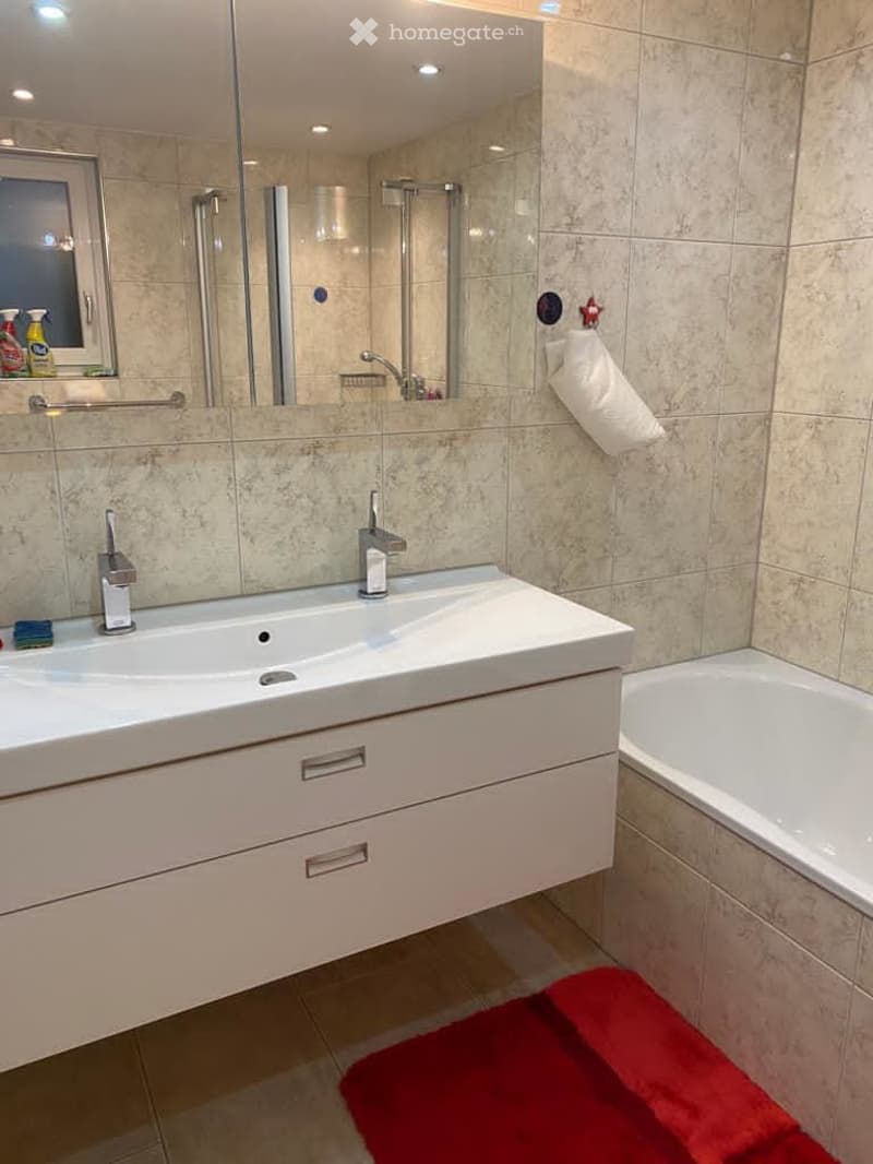 large tiled wall, bathtub, white cabinet with sink, two faucets, mirror, glass shower enclosure, red towel