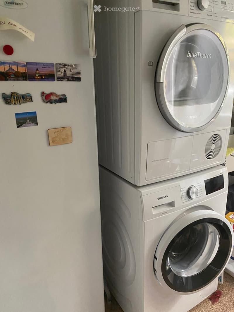 Two stacked white appliances, top is a blueTherm dryer, bottom is a Siemens washing machine, located in a utility room next to a refrigerator.