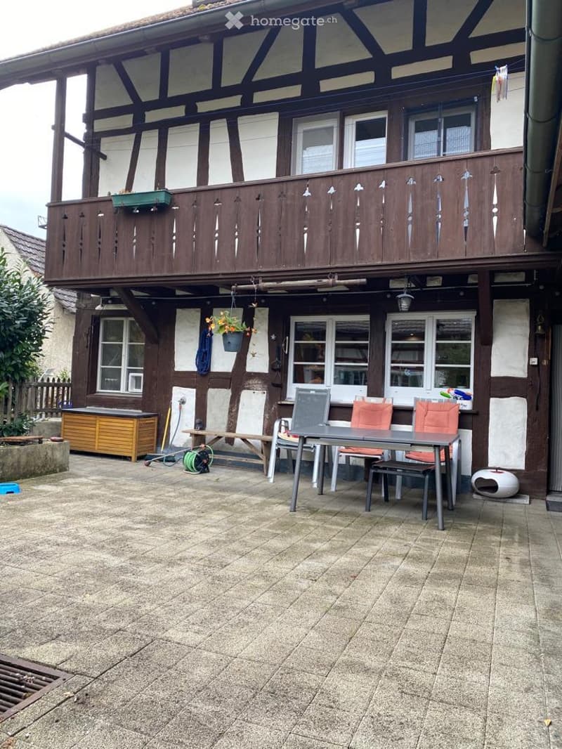 House with brick and timber exterior, balcony, outdoor table with chairs, garden tools, wooden storage box, plants, drainage grate
