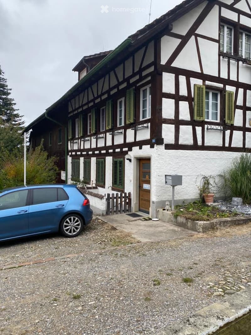A two-story building with brown wooden frame, white walls, and green shutters, a wooden door, a wooden fence in front, a mailbox, a small garden with plants, and a car parked in front.