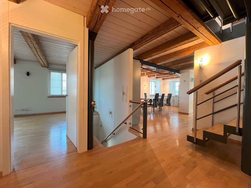 Spacious room, wooden floor, high ceiling with wooden beams, white walls, open staircase to upper floor, glass railings, wall sconces, door leading to another room, electrical outlet on the wall, window with curtains