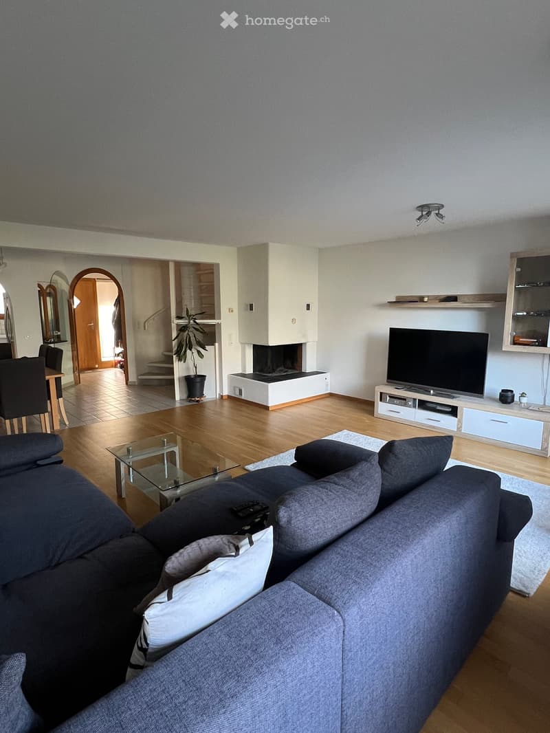 Open living room with couch, TV, and fireplace. Wooden floor, white walls, and a glass table. A plant is in the background. A staircase and a door leading to another room are also visible.