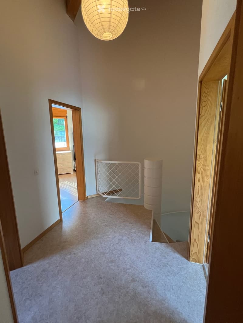 Empty hallway with wooden doors, white walls, carpet, lamp, metal railing, staircase, open door leading to another room with a window.