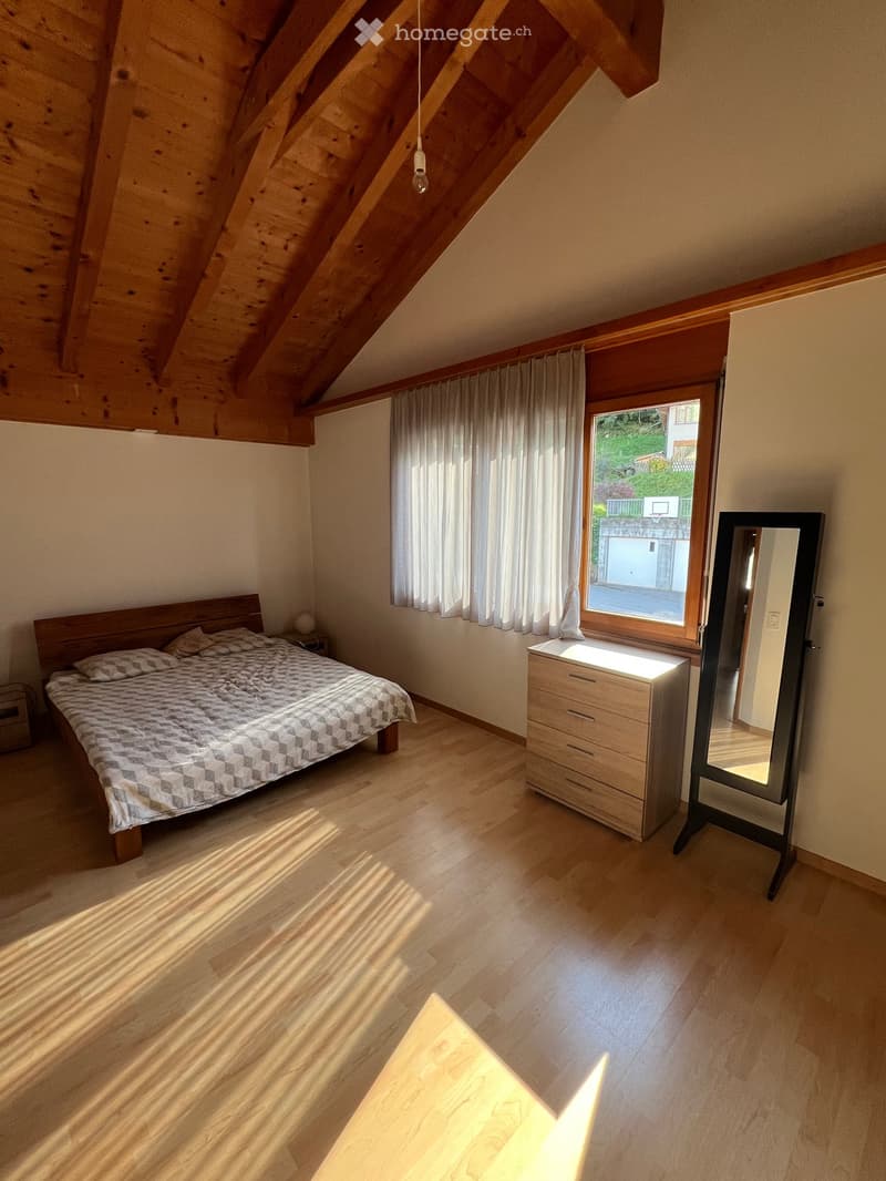 Bedroom with wooden ceiling, bed, pillows, nightstand with lamp, large window with curtains, a wooden dresser, and a mirror. Hardwood floors and bright sunlight.