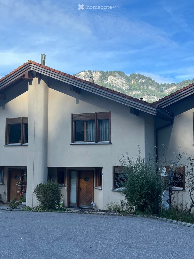 Detached house, beige exterior, brown tiled roof, wooden doors, windows with shutters, surrounded by bushes, front yard with gravel driveway, mountain in the background