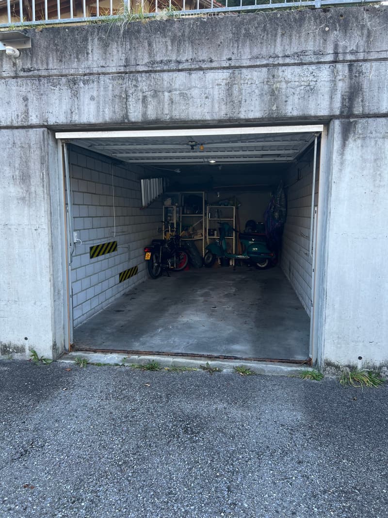 Concrete garage with an open door, two motorcycles parked inside, white walls, metal shelves, and lights mounted on the ceiling.