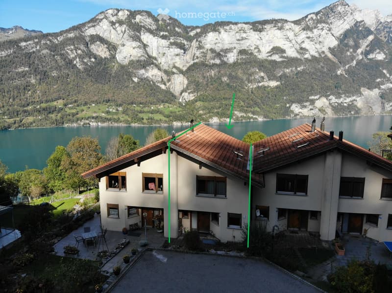 2 story house with brown roof, white walls, multiple windows, lakeside location, large lake in the background, mountains in the distance, green landscape with trees and plants, table and chairs in front of the house, pathway leading to the entrance, small