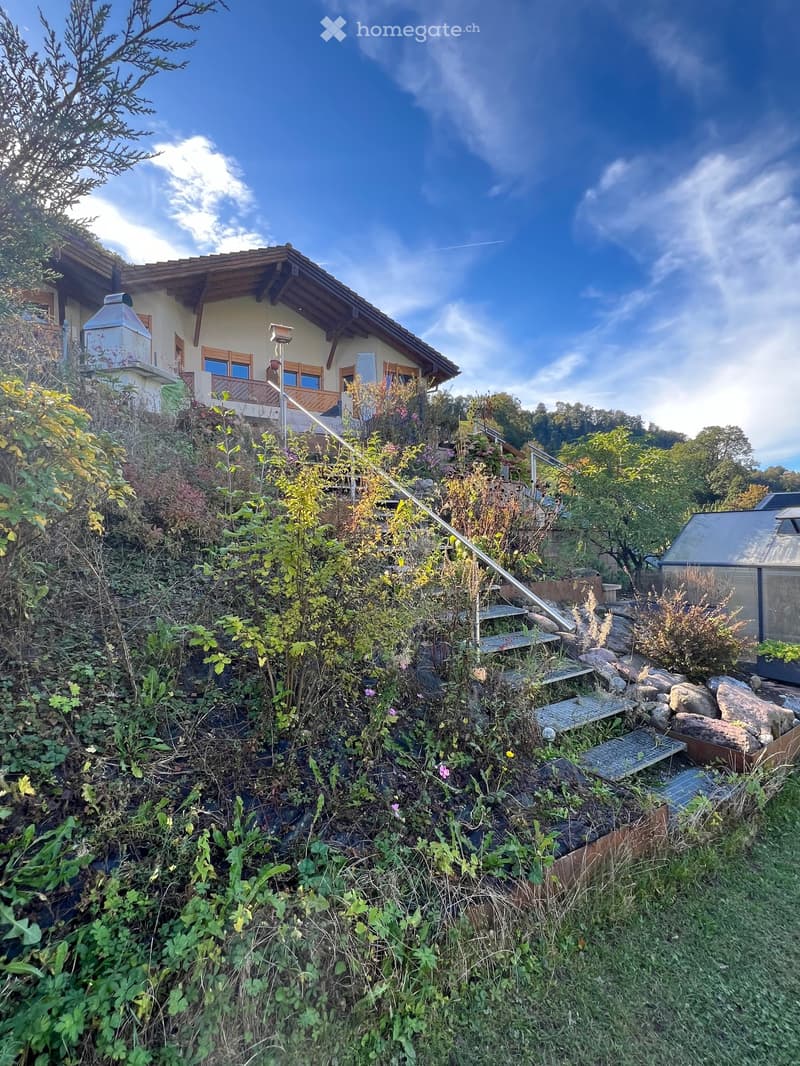 Wooden house with stone steps, brown roof, surrounded by greenery and plants, elevated position, steps leading to the entrance, metal handrail.