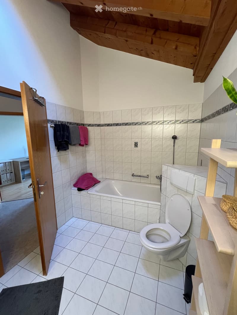 White tiled bathroom with a white bathtub, shower area, toilet, and shelves. White walls and floor, wooden ceiling, open door, towels hanging, and a plant on the shelves.