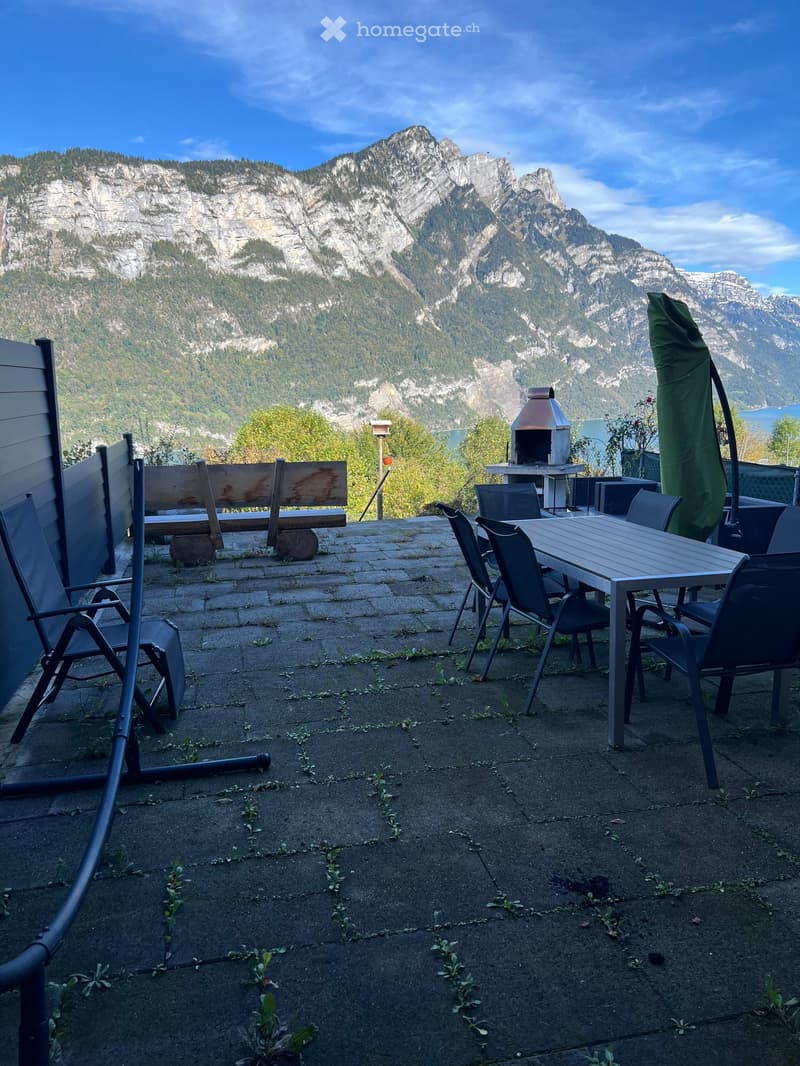 A patio with a dining table, chairs, a bench, a barbecue, and an umbrella; surrounded by plants and mountains with a lake in the distance. The area is paved with stones.