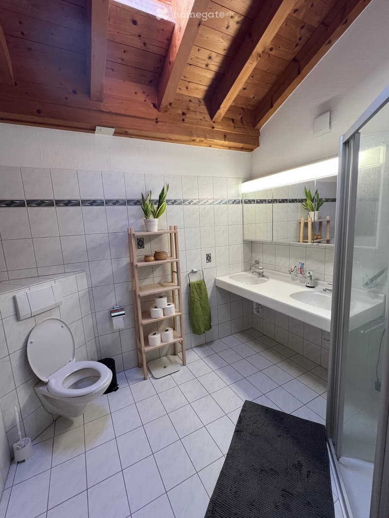 white tiled floor, white tiled walls, wooden ceiling, white toilet, double sink, shower, shelves with plants, towel rack, toilet paper holder