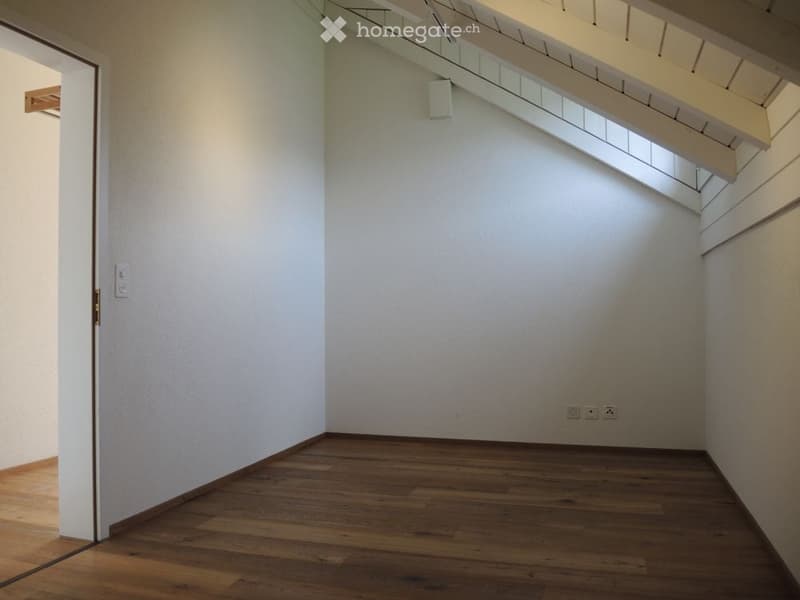 Empty room with wooden floor, white walls, ceiling light, power outlets, slanted roof