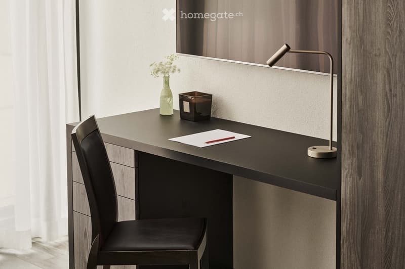 Minimalist home office setup with a dark wooden desk, a black chair, a desk lamp, a vase with flowers, and a small decorative box on the desk. The room has large windows and a wooden wall panel.