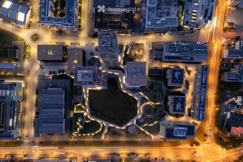 This image shows an aerial view of a city at night. The city skyline is illuminated, with various high-rise buildings and structures visible. There is a large open space or plaza in the center of the image, surrounded by roads and parking areas. The overa