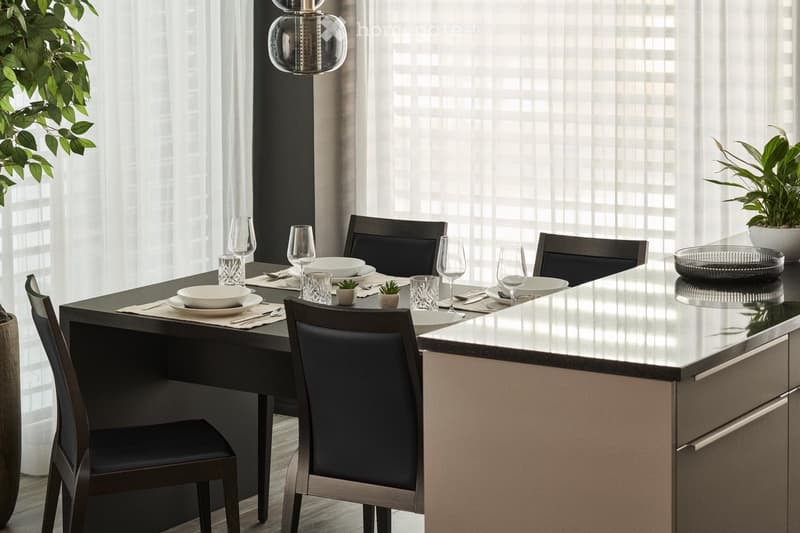 Modern dining room with a rectangular dining table, black leather chairs, a glass pendant light, and a built-in kitchen with a dishwasher. The room has large windows providing natural light and a view of the greenery outside.