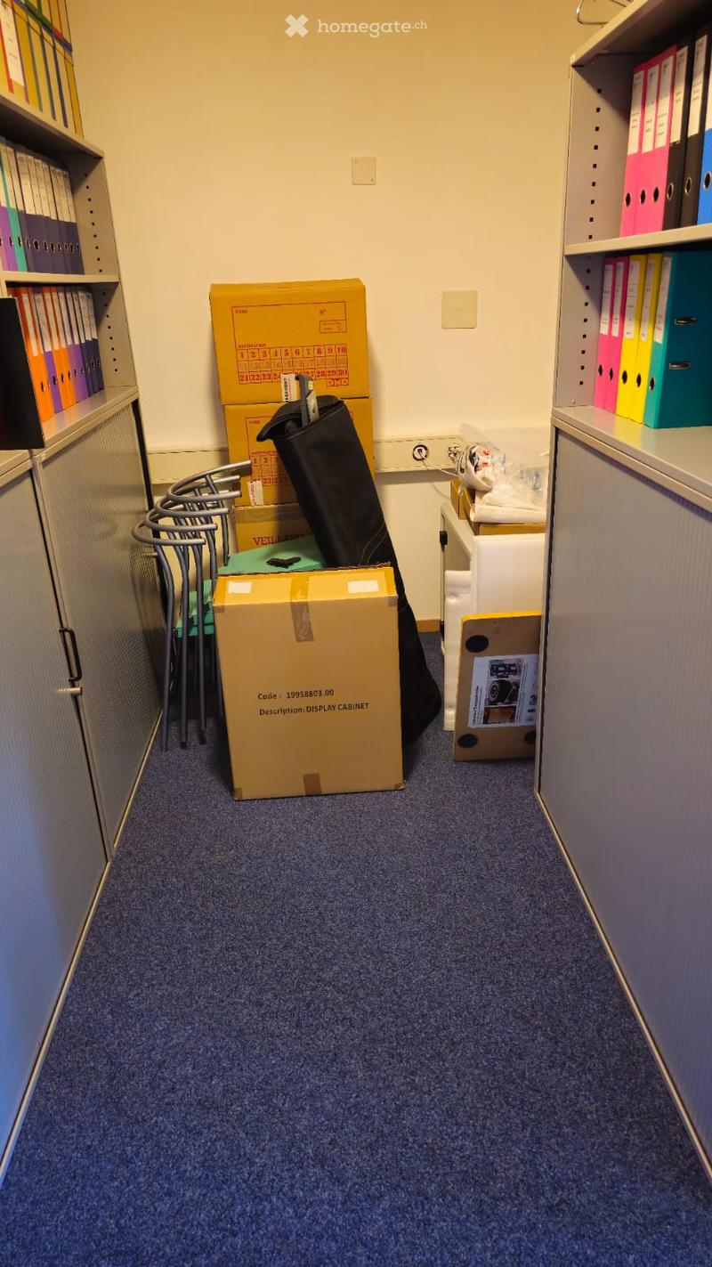 Empty office space with carpet floor, file cabinets, stacked boxes, metal chairs, and miscellaneous office supplies.