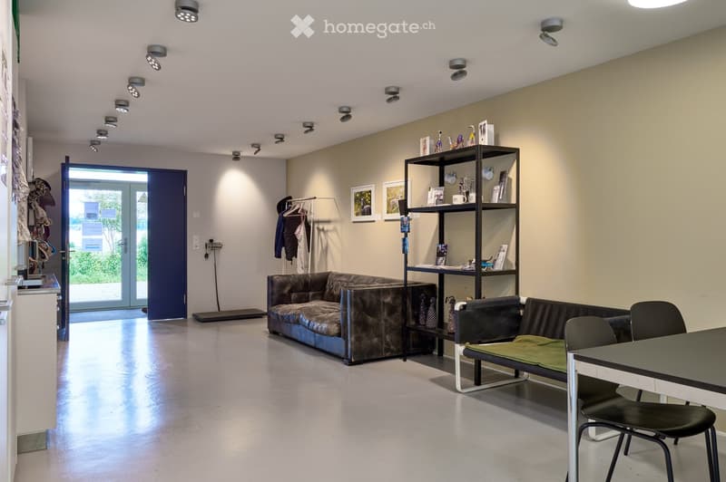 White room with a grey floor, a large sofa, a small couch with cushions, a table, two chairs, a shelf with books and ornaments