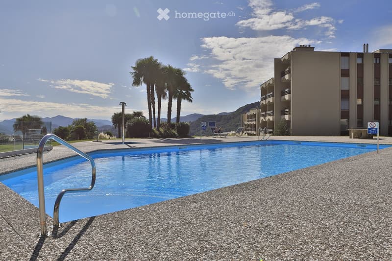 An outdoor swimming pool with a steel handrail, surrounded by gravel flooring. In the distance, there is a building with multiple balconies and a few palm trees. There are mountains in the backdrop, under a blue sky with scattered clouds.