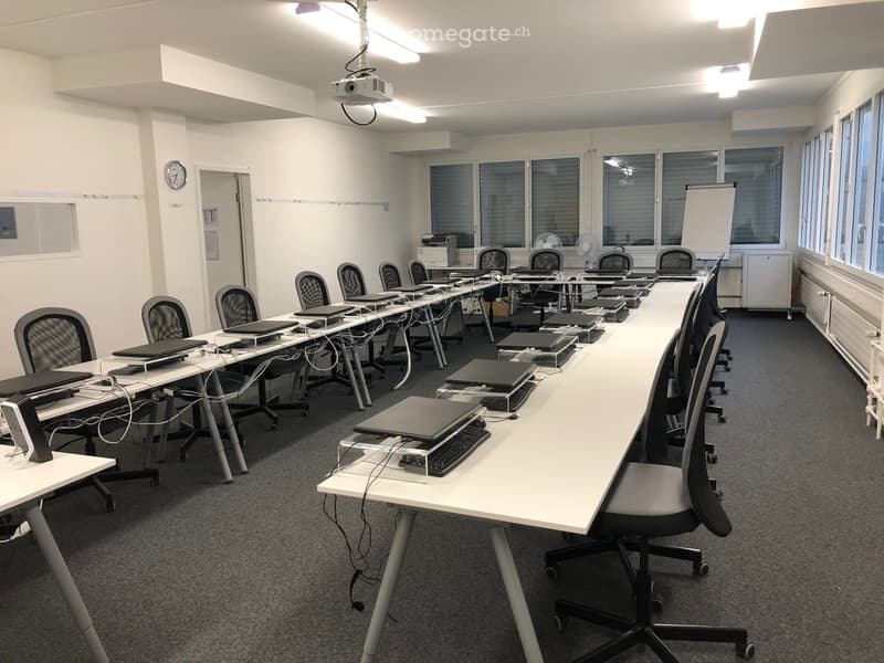 An empty classroom with long tables and chairs. Equipped with laptop stands, monitors, and other office equipment.