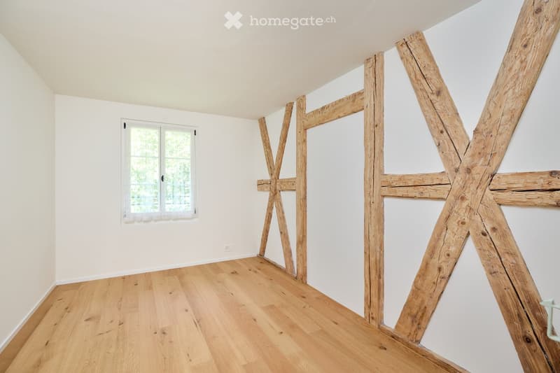 Empty room with wooden beams, hardwood floor, white walls, and a window with white curtains.