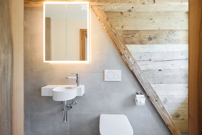 Bathroom with white ceramic sink, chrome faucet, white toilet, gray tiles, wooden ceiling beams, illuminated mirror, toilet paper holder, no visible features