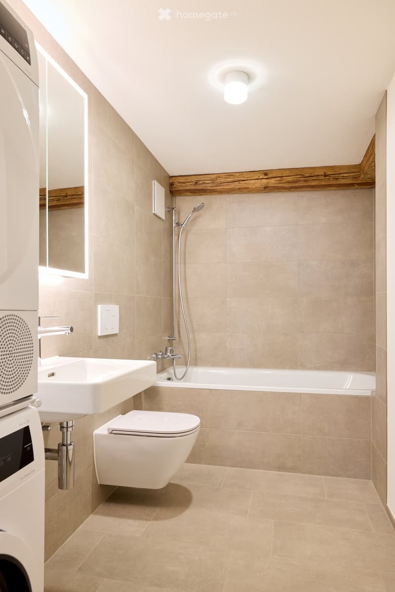 modern bathroom, beige tiles, white sink, shower, toilet, built-in washer, mirror, illuminated wall sconce