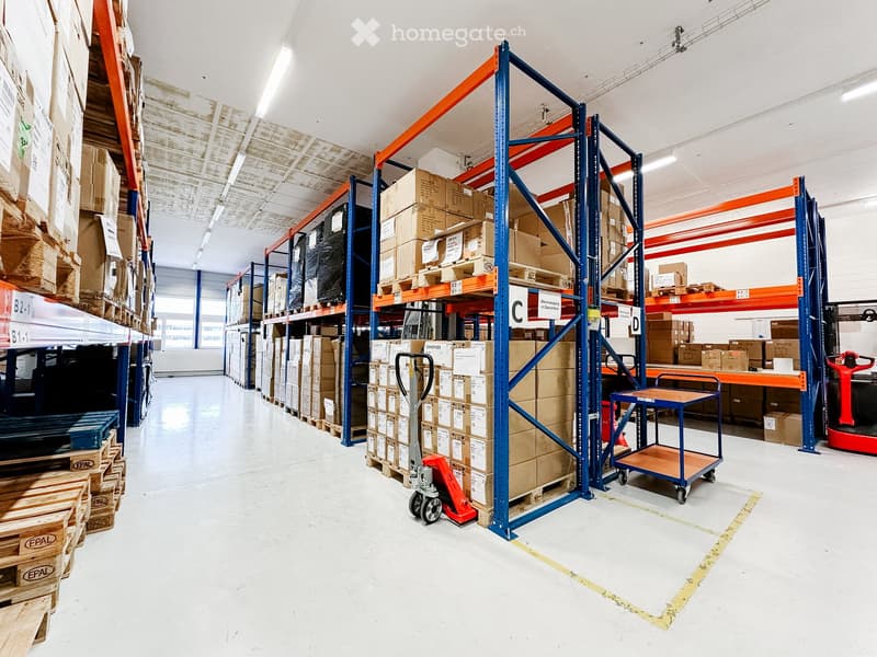 A large warehouse filled with stacked boxes, storage racks, and equipment. There is a cart, a pallet jack, and bright lighting throughout.