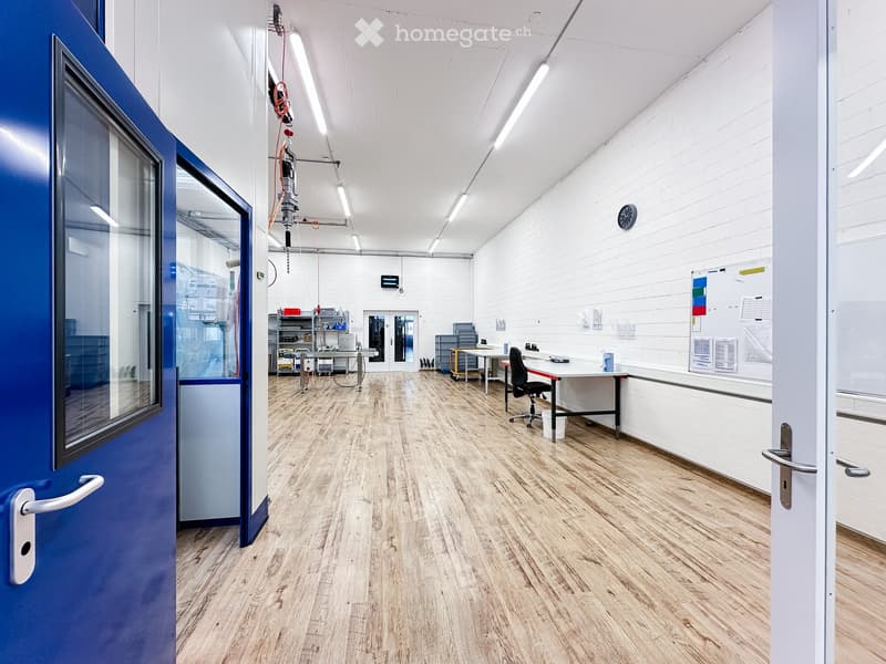 Empty room with wooden floors, white walls, and overhead track lighting. The room has a blue door and a white door on the right side, and contains a table, chair, and some equipment. There is a clock and bulletin board on the wall.