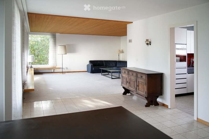 Modern living room with neutral color scheme, wooden ceiling, white tiles on the floor, black sofa, wooden cabinet, glass table, white walls, lamps, white curtains, large window, kitchen with wooden counter, and wooden stools