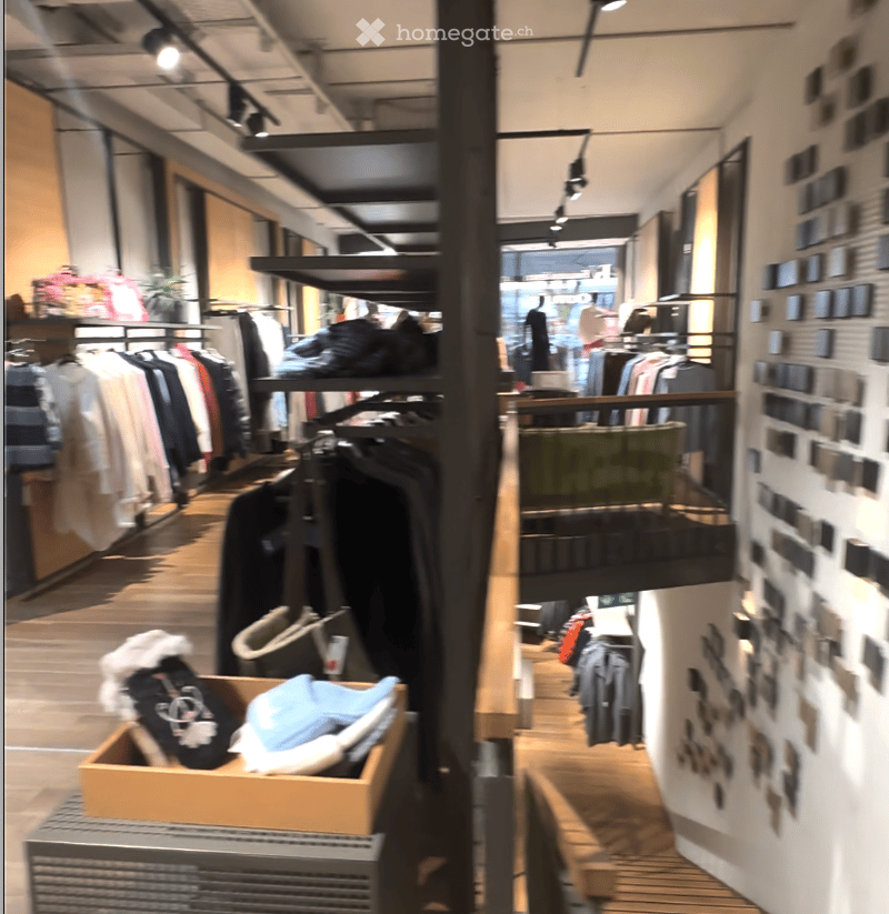 Interior space showing clothing on hangers and on shelves, an entrance at the far end with a mannequin, a wooden desk, and a wooden handrail.