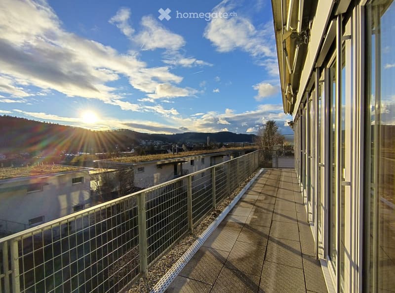 Terrace with green gravel flooring, metal railings, sliding glass doors, green roof on building in background, town and mountains in distance, bright sunshine