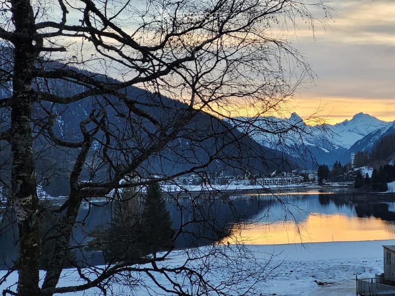 Snowy landscape with a lake, mountains, and a small house