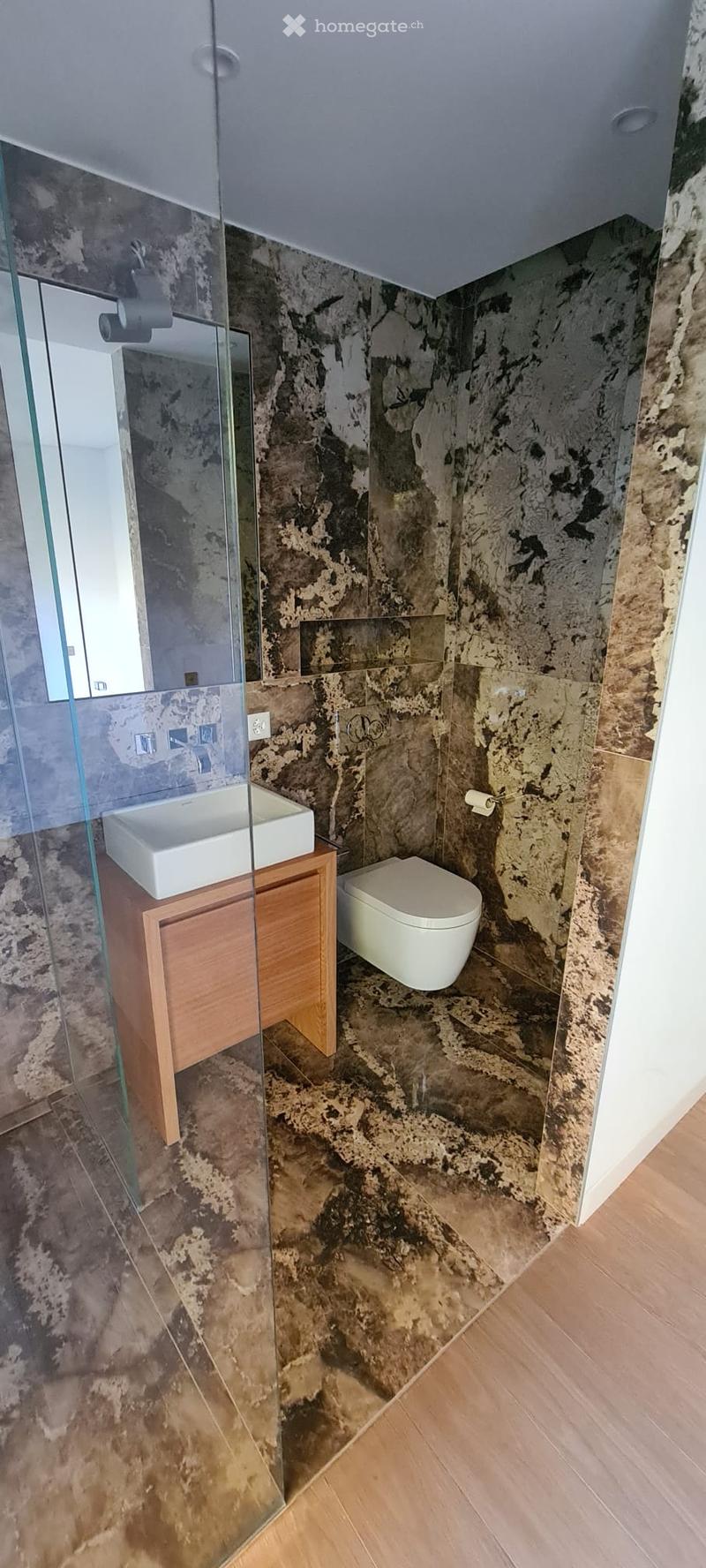 Bathroom with marble walls, glass shower enclosure, white sink, wooden vanity, white toilet, white mirror