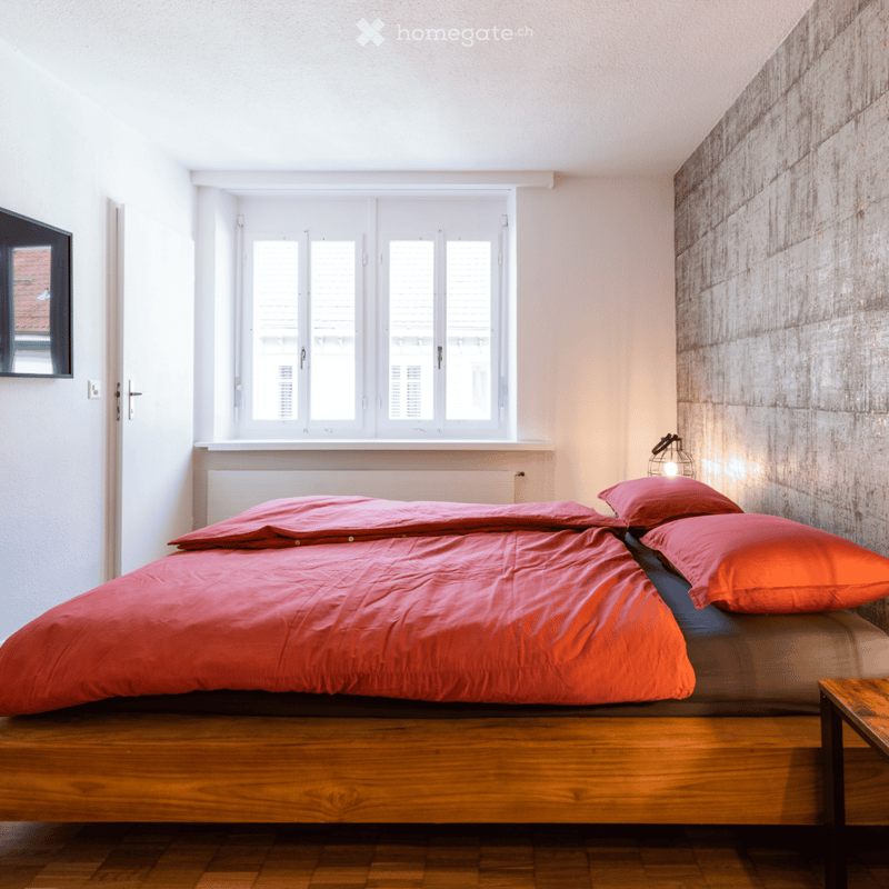 A double bed with red bedding, two red pillows, a wooden table, a window with white shutters