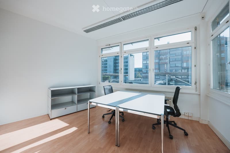 Bright, minimalist office space with wooden flooring, large windows, a white desk, two office chairs, and a grey shelving unit.