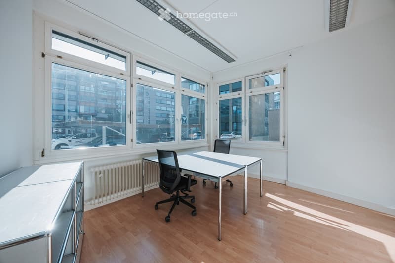 Bright, unfurnished room with large windows, wooden flooring, a white table, and a black office chair.