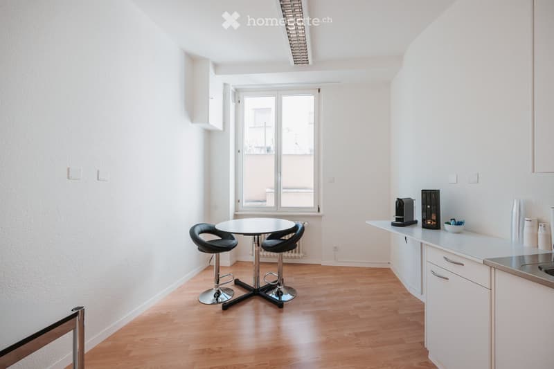 Bright, open-plan room with a small dining area and a kitchen section. Features a round table with two stools, a white kitchen counter with a coffee maker, and built-in cabinets.