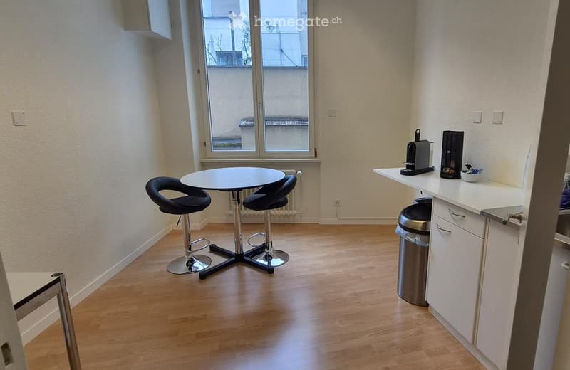 A modern, compact room with a small round table and two black stool chairs, a white kitchen counter with a coffee maker, and a trash bin. The room has light wooden flooring and white walls, with a large window offering a view of the outside.