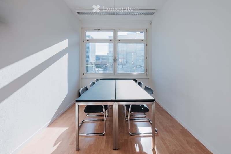 Bright, minimalist room with a long rectangular table and several chairs, large windows letting in natural light, wooden flooring.