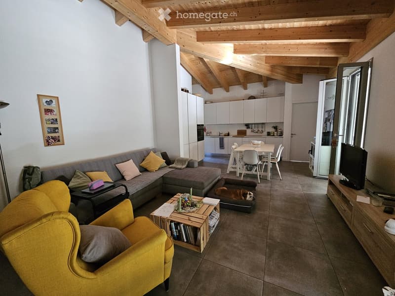 Open-concept living room and kitchen with wooden beamed ceiling, gray sectional sofa, yellow armchair, coffee table, and shelving unit. Tile flooring throughout. Kitchen area with white cabinets and appliances visible.