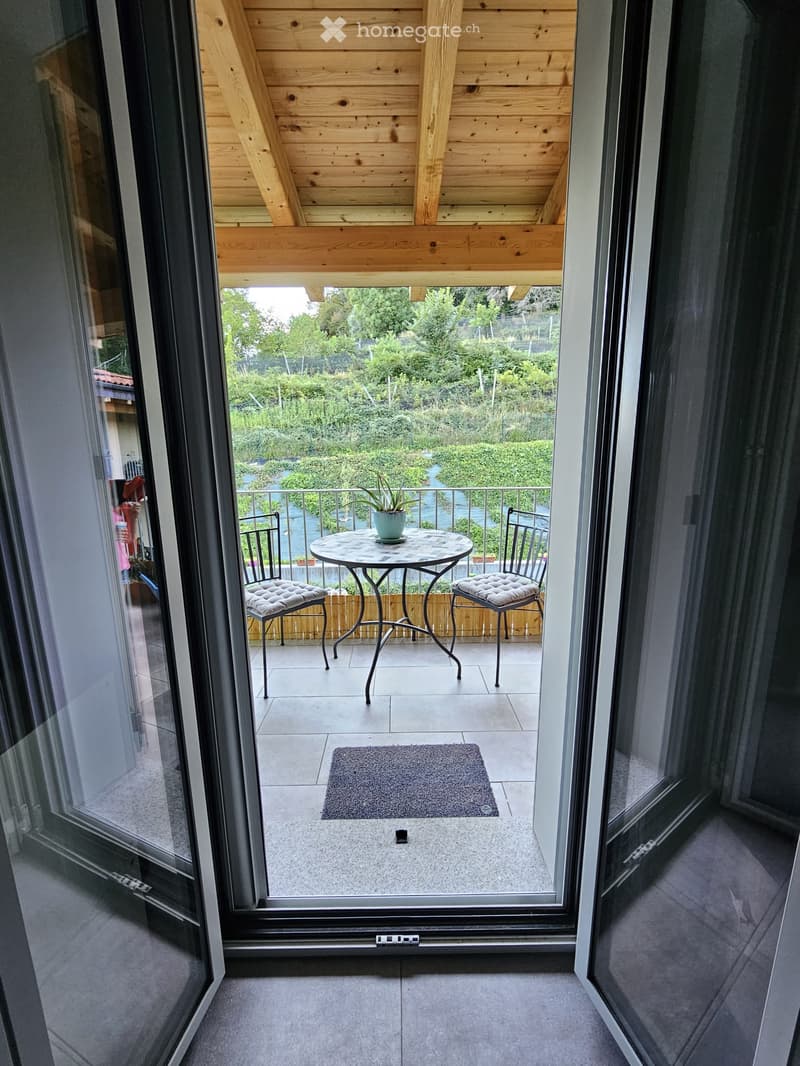 The image shows a balcony or terrace with a wooden ceiling, a round metal table with cushioned chairs, and a view of a lush, green outdoor area through the glass doors.